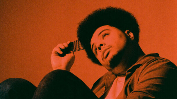Poet Otis Mensah sits, bathed in orange light, combing his hair